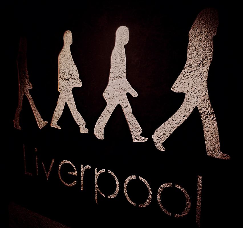 Cartel de Liverpool en la pared: las cuatro siluetas de Abbey Road caminando sobre el nombre, iluminadas con luz cálida